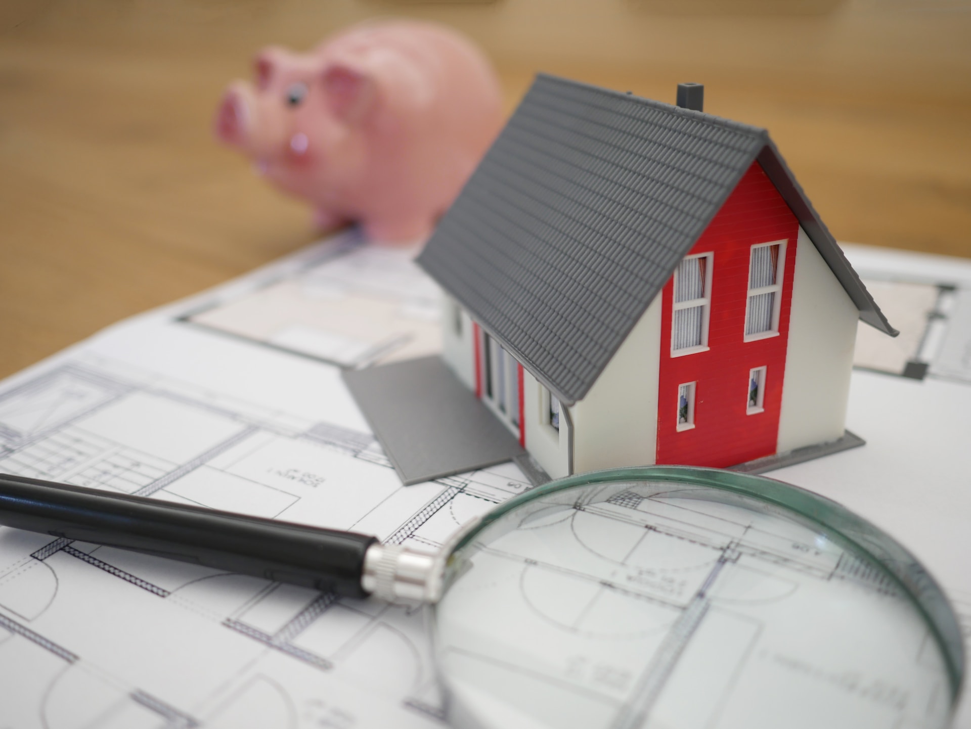 Close up of a model house, sat on a table alongside a magnifying glass and a piggy bank.