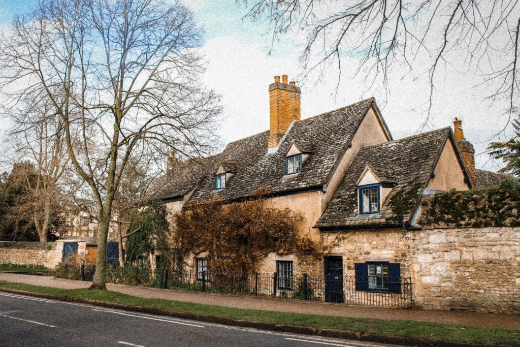 A rural roadside cottage in the British countryside, surrounded by trees and shrubs against the backdrop of a blue sky.