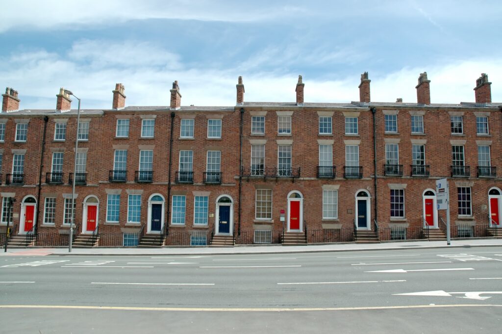 A row of terraced houses viewed from an opposite roadside, against the backdrop of a blue sky.